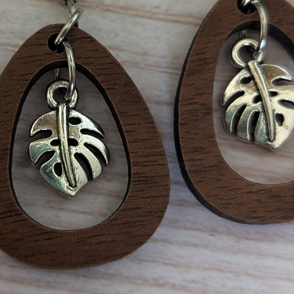 Wooden teardrop-shaped earrings with a silver-toned monstera leaf charm, displayed on a light wood surface.
