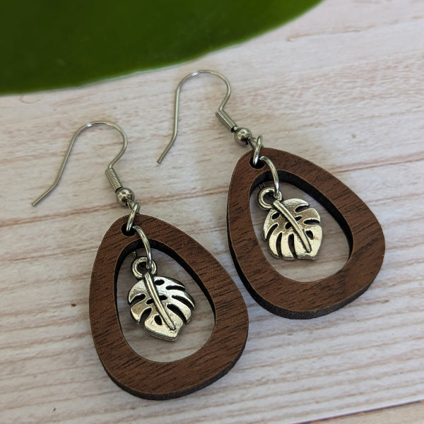 A pair of teardrop-shaped wooden drop earrings with a dark brown wood grain finish, featuring a silver-toned metal leaf charm inside each wooden frame, displayed on a light wood surface.
