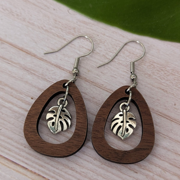 A pair of teardrop-shaped wooden dangle earrings with a silver-toned monstera leaf charm, displayed on a light wood surface with a green leaf in the background.