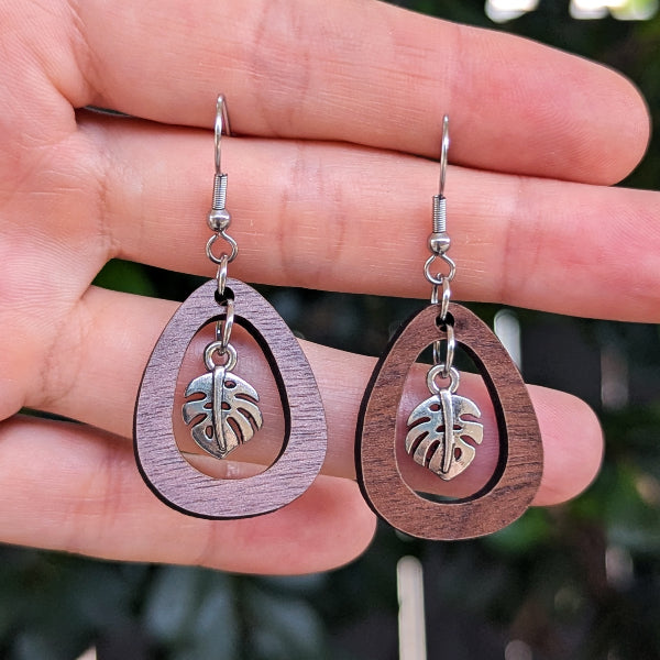 A pair of wooden dangle earrings with teardrop-shaped cutouts and small silver-toned monstera leaf charms, held in a person's hand against a blurred green outdoor background.