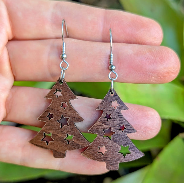 Wooden Christmas tree earrings with star cutouts held between fingers against a green leafy background. Stainless steel hooks.