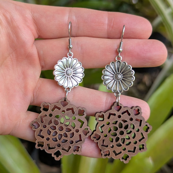 Hand holding a pair of floral and wooden intricate earrings against a natural background. Stainless steel hooks.