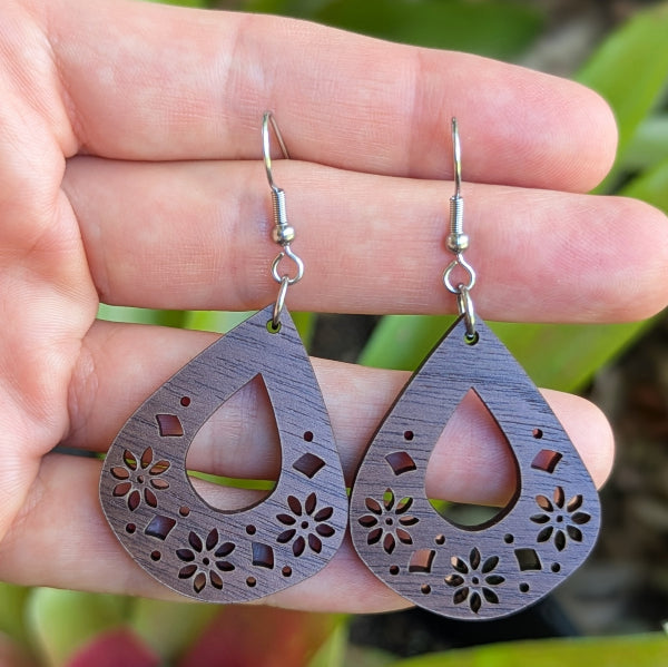 Wooden teardrop earrings with floral cutouts held in a hand against a natural background. Stainless steel hooks.