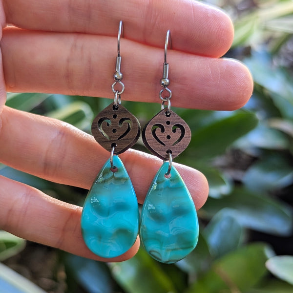 Resin turquoise coloured teardrop earrings with wooden accents on a light wood surface. Held between fingers. Stainless steel hooks.