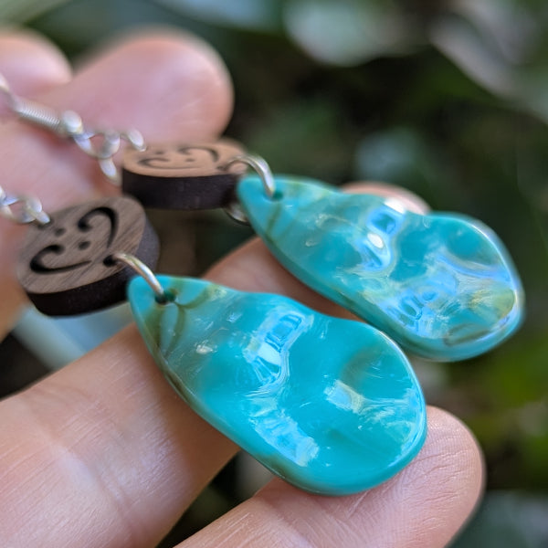 Resin turquoise coloured teardrop earrings with wooden accents on a light wood surface. Held in finger tips. Stainless steel hooks.