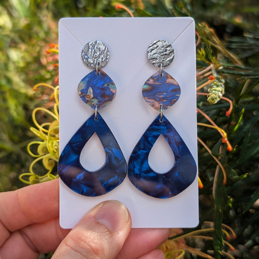 A pair of drop earrings displayed on a white card, held by a hand against a blurred outdoor background of green foliage and yellow flowers. The earrings feature textured silver studs, iridescent blue teardrop beads, and larger marbled blue teardrop pendants.