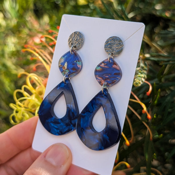 A pair of dangling earrings displayed on a white card, held by a hand against a blurred outdoor background of green foliage and yellow flowers. The earrings feature textured silver-tone studs, iridescent blue and purple marbled beads, and large dark blue marbled teardrop drops with cutout centers.