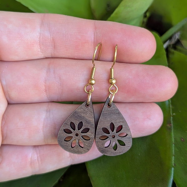 Wooden teardrop earrings with floral cutout held between fingers against a blurred green background. Gold plated stainless steel hooks.
