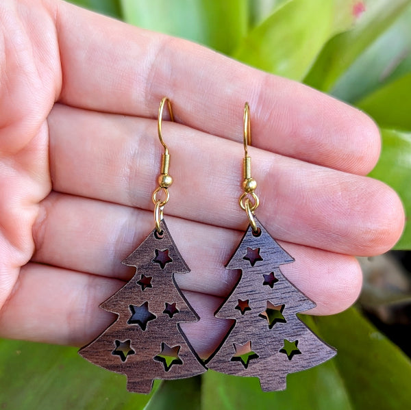 Wooden Christmas tree earrings with gold hooks held in a hand against a green leafy background.