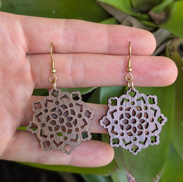 Wooden earrings with intricate cut-out designs held in a hand against a green leafy background. Gold plated stainless steel hooks.