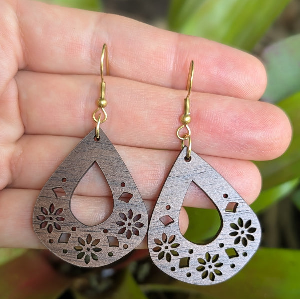 Wooden teardrop earrings with floral cutouts held in a hand against a natural background. Gold plated stainless steel hooks.