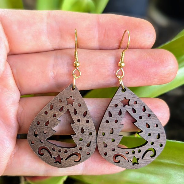 Wooden teardrop earrings with star and tree cutouts held between fingers against a blurred green background. Gold plated stainless steel hooks.