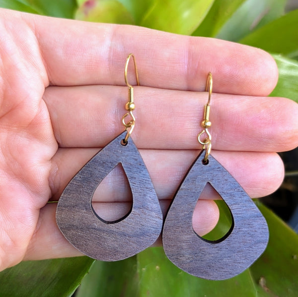 Wooden teardrop earrings held in a hand with a green leafy background. Gold plated stainless steel hooks.