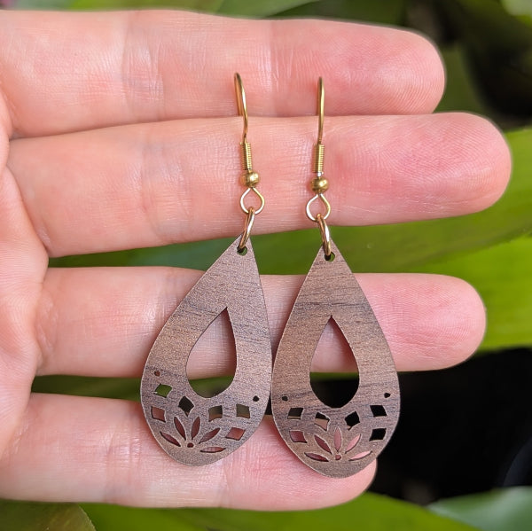 Wooden teardrop earrings with floral cutouts held on a hand. Gold plated stainless steel hooks.
