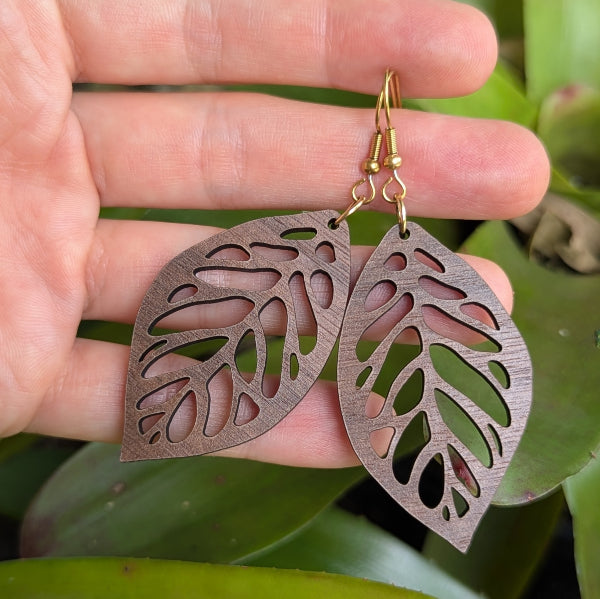 Wooden leaf-shaped earrings with gold tone hooks held in a hand with a green leafy background