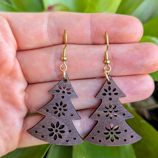 Wooden Christmas tree-shaped earrings with floral cutouts held in a hand against a green leafy background. Gold plated stainless steel hooks.