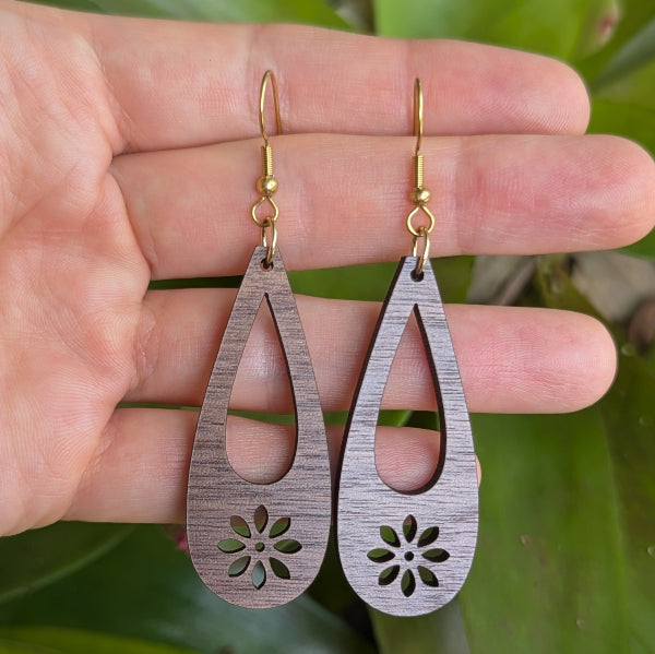 Wooden teardrop earrings with floral cutouts held between fingers against a natural background. Gold plated stainless steel hooks.