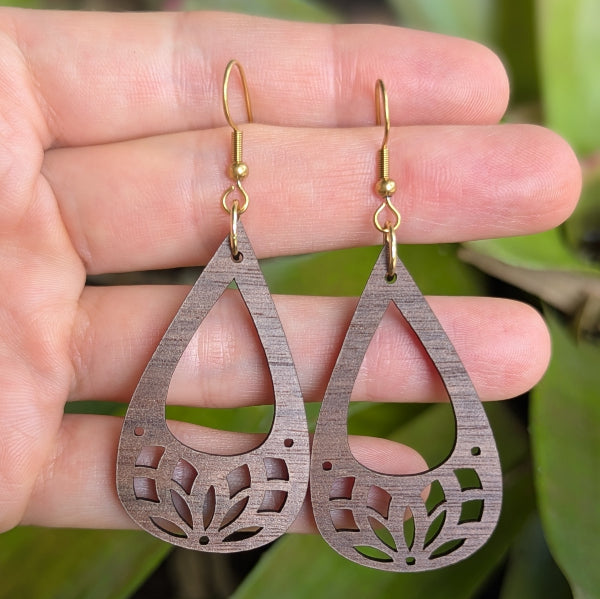 Wooden teardrop earrings with intricate cut-out designs held between fingers against a green leafy background. Gold plated stainless steel hooks.