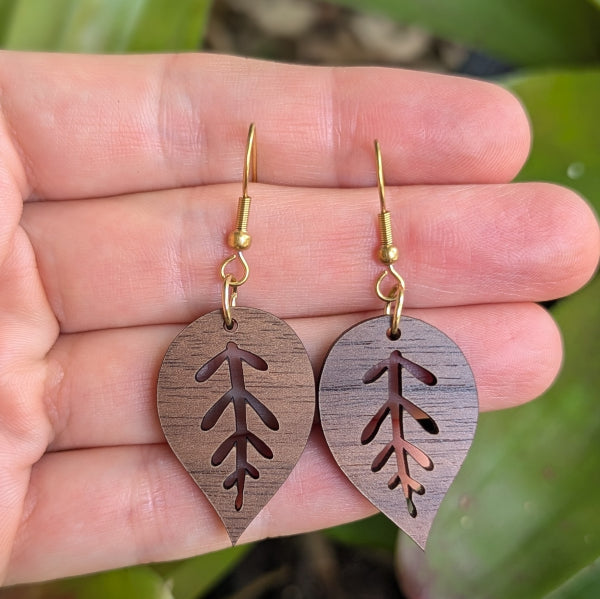 Wooden leaf-shaped earrings with gold plated hooks held between fingers against a natural background.