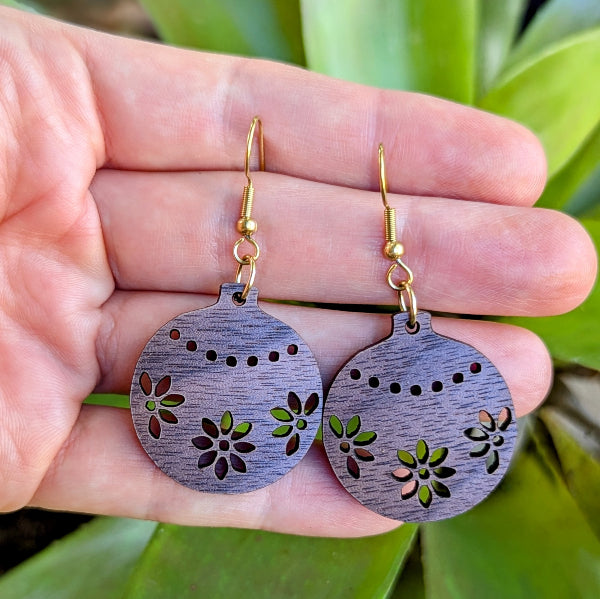 Wooden Christmas bauble earrings with floral patterns held in a hand against a green leafy background. Gold plated stainless steel hooks.