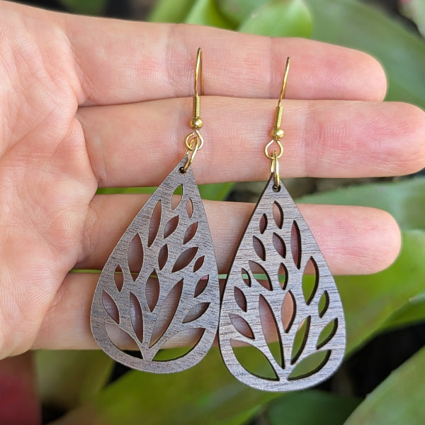 Wooden tear drop earrings held on a with green leaves in the background. Gold plated stainless steel hooks.