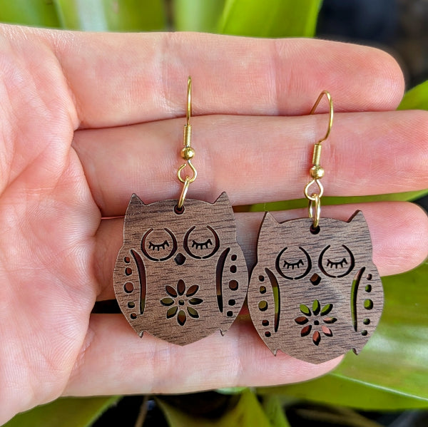 Wooden owl earrings held between fingers with a blurred natural background. Gold plated stainless steel hooks.