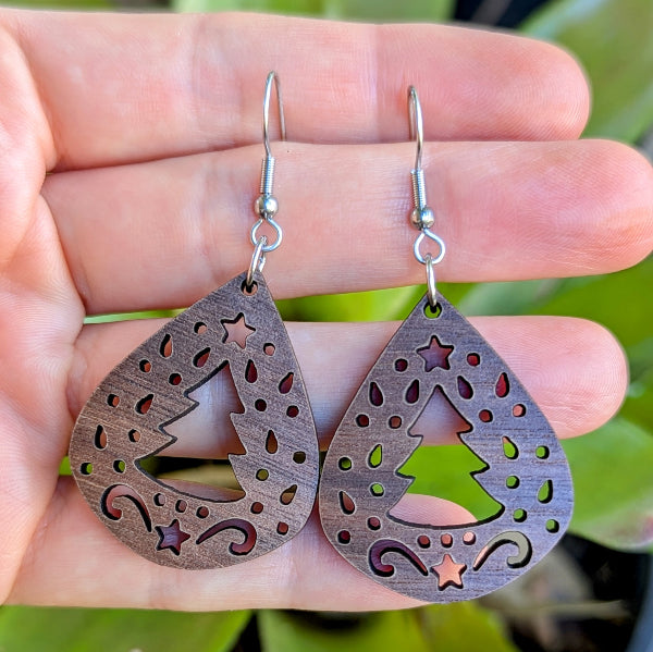 Wooden teardrop earrings with star and tree cutouts held between fingers against a blurred green background. Stainless steel hooks.