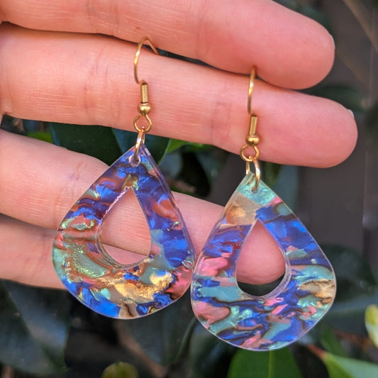 Pair of marbled blue, pink and gold acrylic earrings held in hand. Gold tone hooks, leafy background.