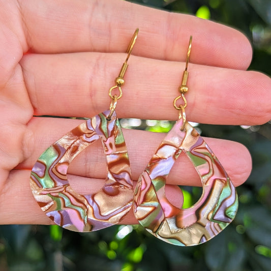 A pair of teardrop-shaped dangle earrings with a marbled iridescent shell-like pattern in pink, green, gold, and brown, hanging from gold fishhook wires and held in a person's hand against a blurred green outdoor background.