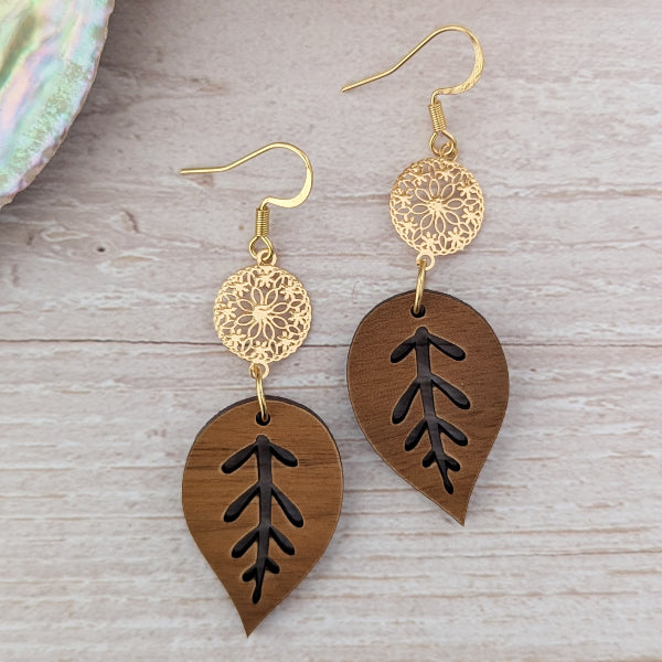 Wooden leaf-shaped earrings with gold floral components on a wooden surface