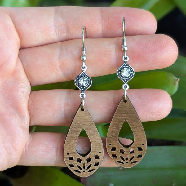 Wooden teardrop earrings with silver toned connectors with crystals, stainless steel hooks held on a hand.