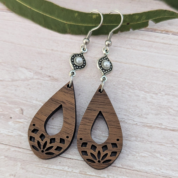 A pair of teardrop-shaped wooden dangle earrings with laser-cut floral patterns, silver-tone hooks, and small pearl-like accents, displayed on a light wood surface with a green leaf.