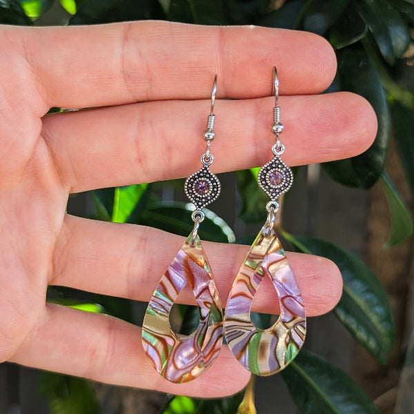 A pair of teardrop-shaped dangle earrings with iridescent acrylic showing pink, purple, green, and gold swirls, suspended from silver hooks and held in a hand against a background of green leaves.