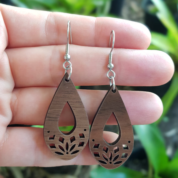 Wooden teardrop earrings with floral cutouts held on a hand. Stainless steel hooks.