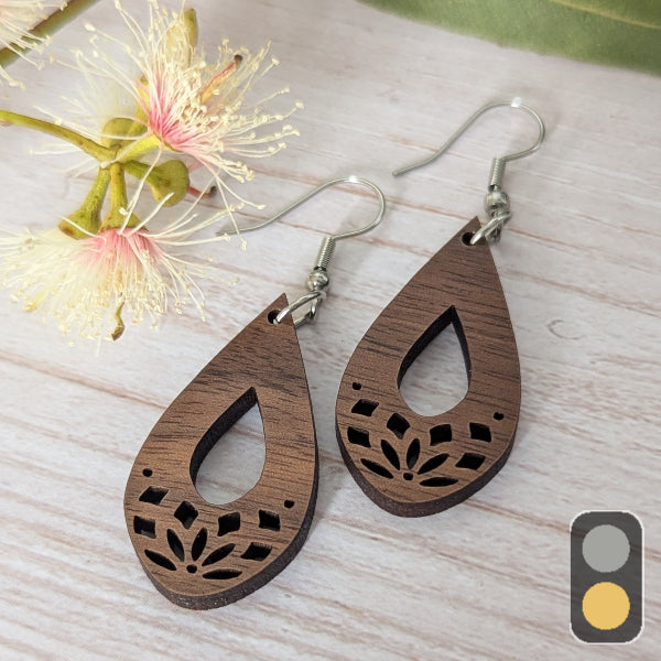 Wooden teardrop earrings with floral cutouts on a light wooden surface with flowers. Stainless steel hooks.