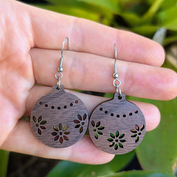 Wooden Christmas bauble earrings with floral patterns held in a hand against a green leafy background. Stainless steel hooks.