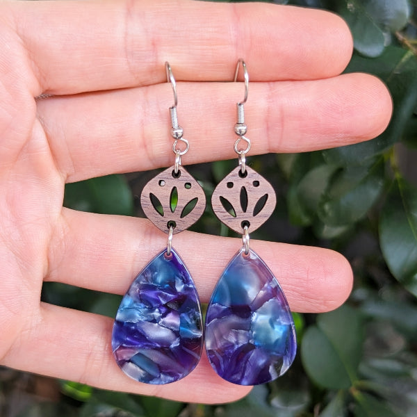Teardrop-shaped earrings with purple and blue marbled design held between fingers against a blurred green background.