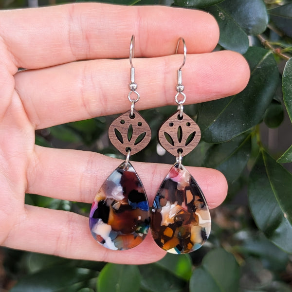 Teardrop-shaped earrings with multicolored design held between fingers against a leafy background