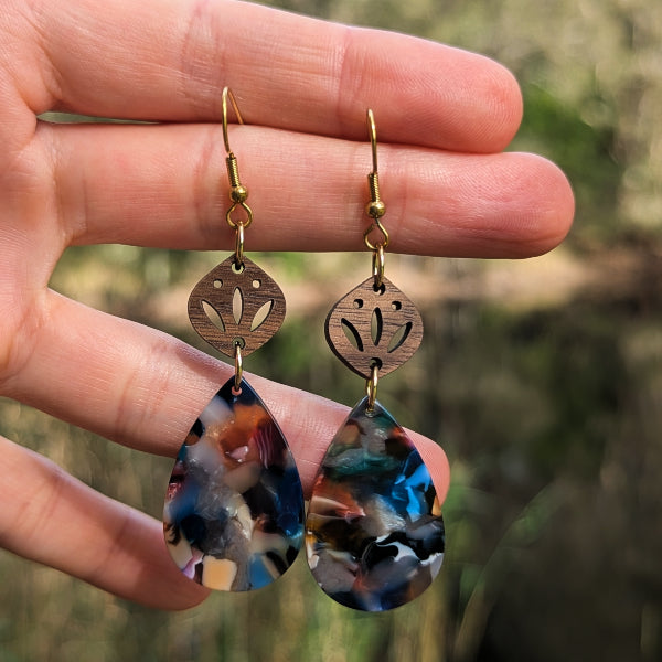 A pair of handmade teardrop earrings held in a hand. The earrings feature a bronze-toned metal top with a cutout floral design and a dangling marbled resin teardrop charm in blue, black, white, and orange.