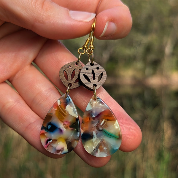 A close-up of a hand holding a pair of dangling earrings outdoors. The earrings have gold-tone fishhook wires, oval wooden top pieces with cutout leaf-like patterns, and teardrop-shaped resin drops containing multicolored natural debris.