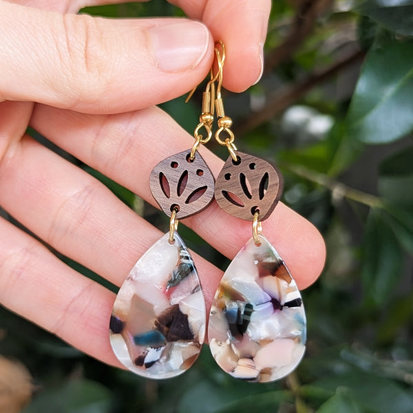 Hand holding a pair of earrings with teardrop-shaped drops and leaf-like wooden components against a blurred green background.