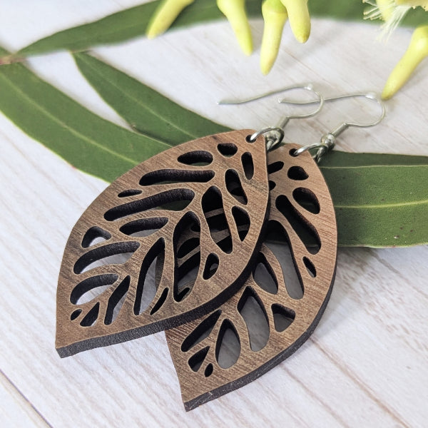 Wooden leaf-shaped earrings on a white surface with green leaves in the background