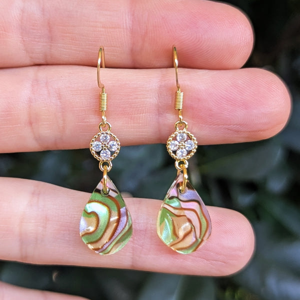 A pair of gold-tone dangle earrings with iridescent green, gold, and purple teardrop charms, held between fingers against a blurred dark green background.