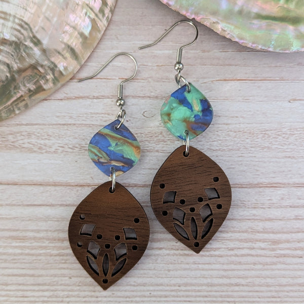 A pair of handmade dangle earrings featuring iridescent blue, green, and gold acrylic connectors and laser cut dark brown wooden leaf-shaped drops, displayed on a light wood background with a shell prop.