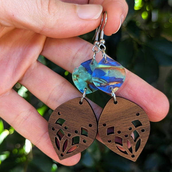 A close-up of a hand holding a pair of dangling earrings against a blurred green outdoor background. The earrings have silver hook wires, round iridescent blue-green connectors, and brown wooden leaf-shaped drops with laser-cut geometric and floral patterns.
