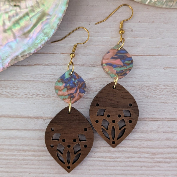 A pair of handmade dangle earrings featuring dark brown laser-cut wooden teardrop pendants and iridescent blue, pink, and green acrylic disc charms on gold-tone hook wires, displayed on a light wood background.
