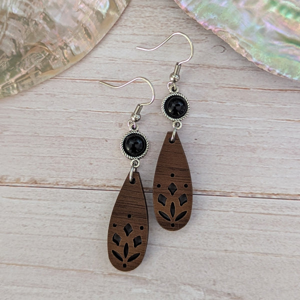 A pair of bohemian-style wooden dangle earrings with laser-cut floral patterns, black stones, and silver-tone hook wires, displayed on a light wood surface.