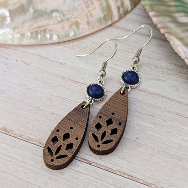 A pair of bohemian-style wooden dangle earrings with laser-cut floral patterns, dark blue oval stones, and silver-tone hook wires, displayed on a light wood surface.