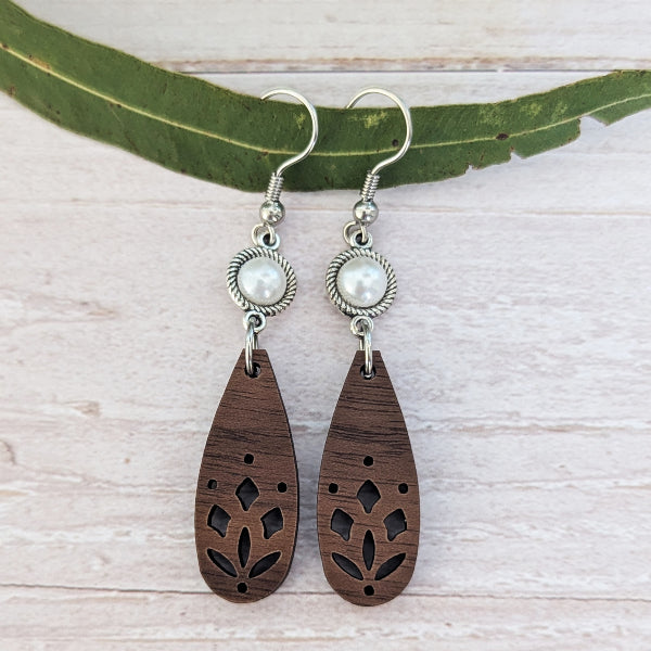 A pair of handmade teardrop wooden dangle earrings with laser-cut floral patterns, featuring a silver-toned textured bezel holding a white faux pearl, hanging from silver fishhook wires. The earrings are displayed on a light-colored wooden surface with a green leaf prop.