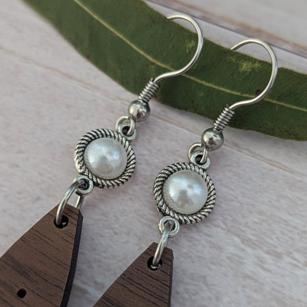 A pair of handmade dangle earrings featuring silver-toned twisted wire circles with white faux-pearl beads, hanging from silver fishhook wires, and finished with dark brown wooden teardrop pendants. The earrings are displayed on a light wood surface with a green leaf prop.